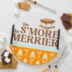 A round sign with “the S’more the Merrier” in brown text and an illustrated s’more. Surrounding it are graham crackers, chocolate, marshmallows, and roasting forks on a white surface.