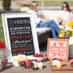 A festive outdoor table displays patriotic decorations, snacks, and fruit. A framed sign with "America!" lyrics and a "1776" sign are featured. Two women sit blurred in the background, with sunflowers adding a cheerful touch.