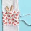 A white apron with a pocket patterned with watermelon slices holds two wooden kitchen utensils, set on a light blue background.