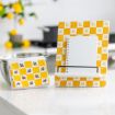 A yellow and white checkered recipe stand with bee patterns holds a blank spiral notebook. Beside it is a matching tea towel on a bowl. The background shows a kitchen with lemons and a pot on the stove.