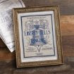 A framed vintage-style print with ornate gold edges displays sheet music titled "The Liberty Bells Are Ringing," featuring decorative blue text and flourishes on a rustic wooden surface with another copy partially visible nearby.