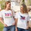 Two women smiling at each other outdoors, both wearing white t-shirts with American flag designs. One shirt has multiple flags on a clothesline, and the other has a single waving flag with the words "God Bless.