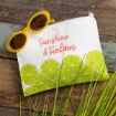 A white zipper pouch with green lime slices and the words "Sunshine & Tanlines" in pink script, next to yellow sunglasses on a rustic wooden surface with green grass blades in the corner.