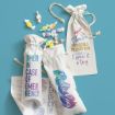 Three white drawstring bags with colorful text, surrounded by pastel wrapped candies. The bags read: “OPEN IN CASE OF EMERGENCY,” “Goodie Bag,” and “You’re impossible to shop for ~but I gave it a try.”.