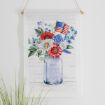 A wall hanging featuring a blue mason jar labeled "EST. 1776," filled with red, white, and blue flowers and a small American flag. Greenery accents surround the jar, and a plant is visible in the lower left corner.