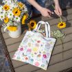A person trims yellow flowers at an outdoor table with a bouquet of daisies and sunflowers in a white vase. A colorful tote bag decorated with garden-themed illustrations rests nearby. Gardening tools and flowers are on the table.