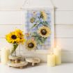 A decorative arrangement features sunflowers in a glass vase with three pillar candles and round wooden coasters. A hanging cloth with sunflower artwork is displayed on a white shiplap wall in the background.