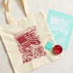 A beige tote bag with a red American flag design lies on a light surface next to a blue sheet with the same flag print, a small turquoise box, and a tin with red ink.