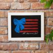 A framed artwork hangs on a brick wall, featuring red stripes, a large blue bow, and the words "All American Girl" in white script. Green leaves partially frame the image on the right side.