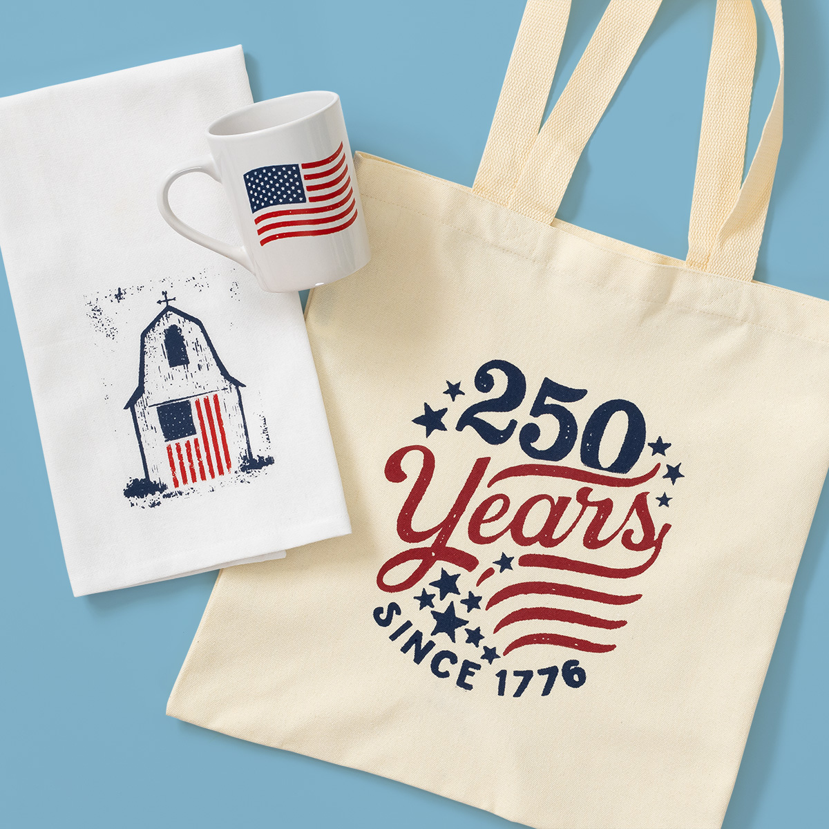 A cream tote bag reads "250 Years Since 1776" with stars and a waving flag. Next to it are a white mug with an American flag and a white tea towel featuring a church with a flag design on the door. Blue background.