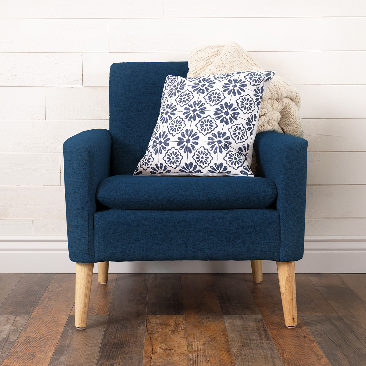 A modern blue armchair with wooden legs sits on a wooden floor. A white cushion with a navy floral pattern and a cream knit blanket are placed on the chair. White shiplap wall is in the background.