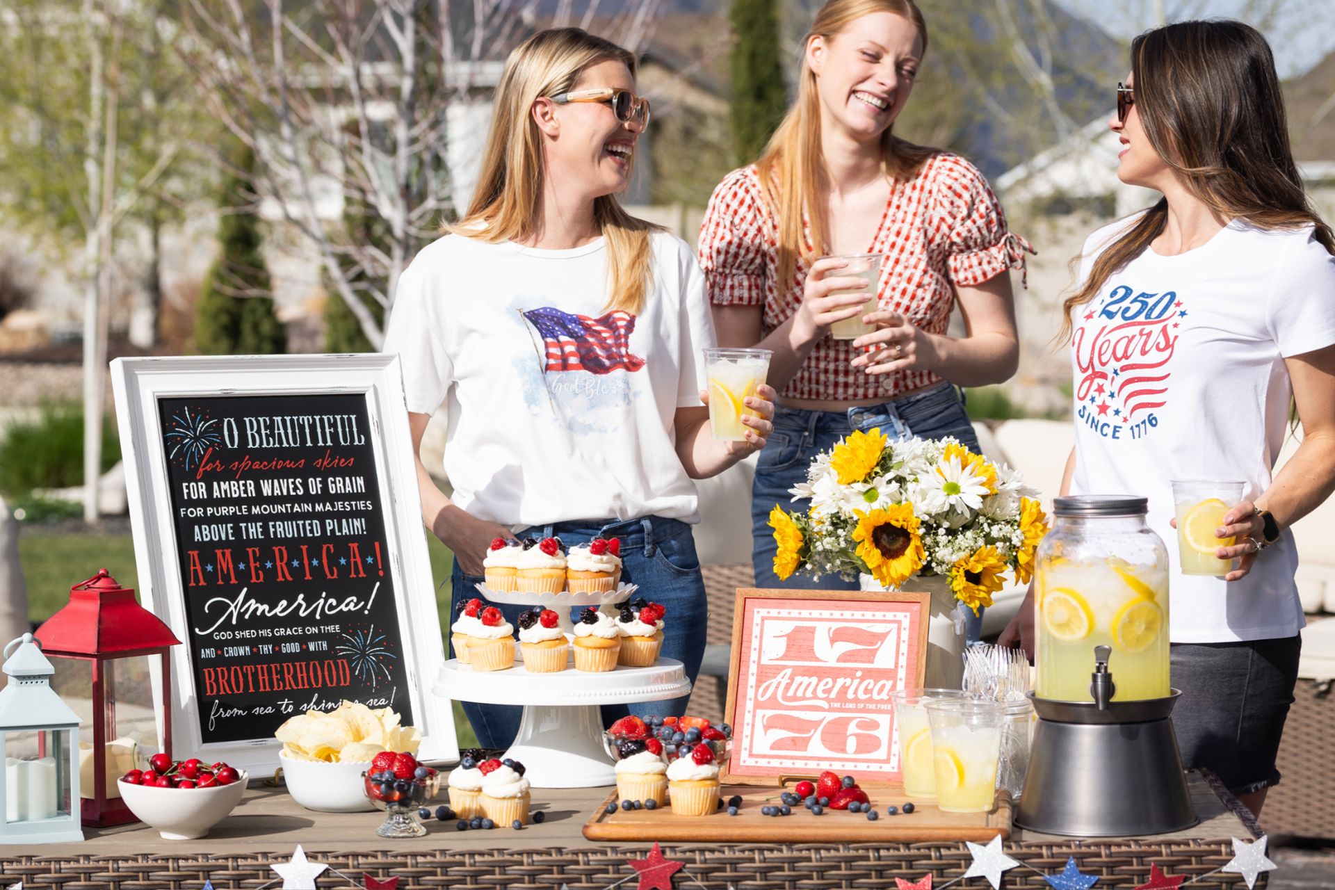 Three women laugh and enjoy lemonade at a sunny outdoor table decorated with patriotic items, cupcakes, sunflowers, and a chalkboard sign celebrating America’s independence. A pitcher of lemonade and festive decorations are displayed.