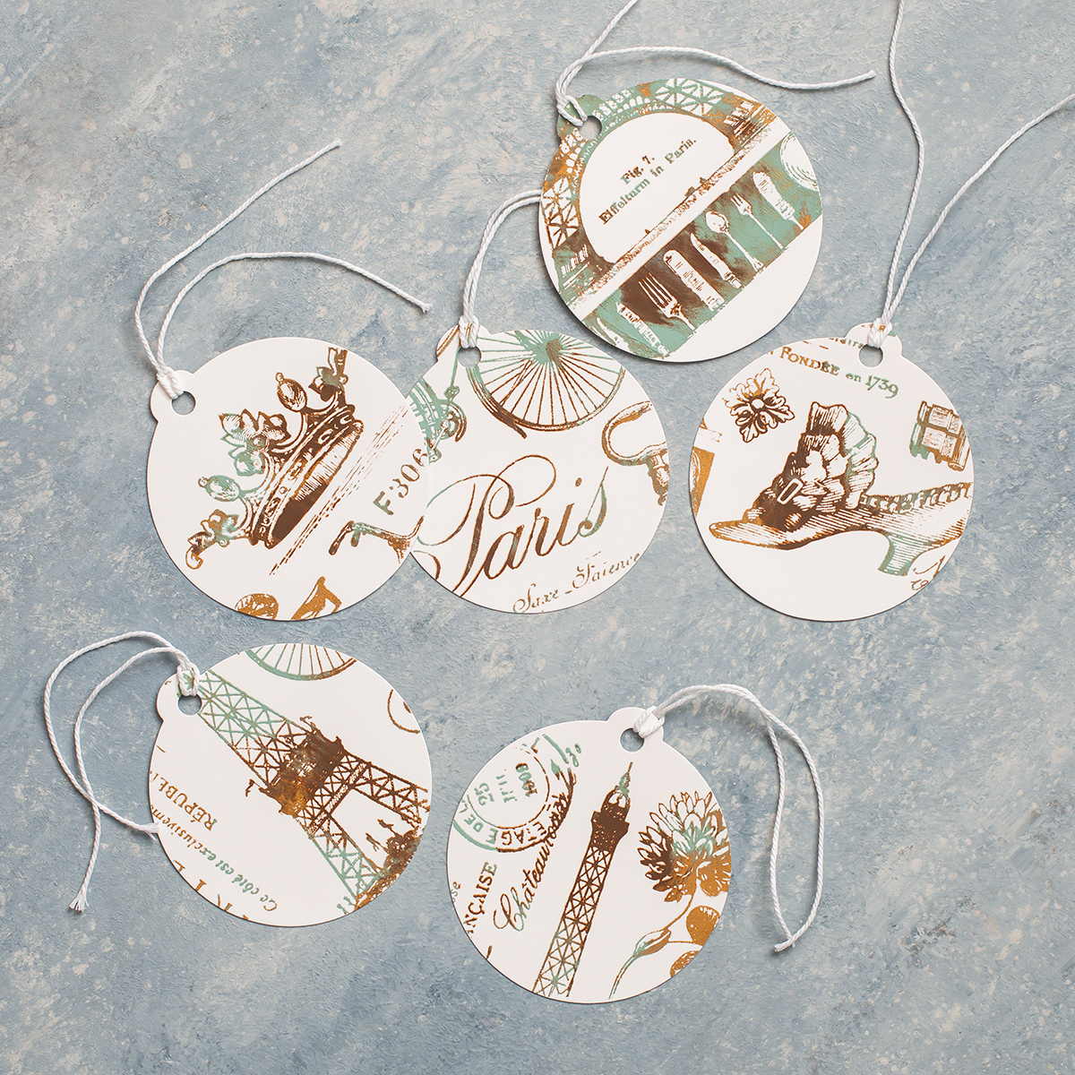 Six round gift tags with Paris-themed illustrations and writing, featuring images of the Eiffel Tower, flowers, shoes, and French text, are arranged on a light blue textured surface. Each tag has a white string.