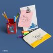 A pencil holder, notepad, and pouch with Stitch designs are arranged on a blue surface with pens and pencils. A pink note on the notepad says “Party Friday 7:00!” The items feature Disney’s Stitch character.