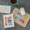 A notebook with Lilo & Stitch characters, a mouse on a colorful mousepad featuring Stitch by a van with "Aloha" text, a keyboard, and a mug of coffee on a gray desk.