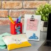 A desk with Stitch-themed stationery: a pencil holder with pens, a white pouch with Stitch’s drawing, and a white clipboard with a note reading “Party Friday @ 7:00!”. A laptop and a potted plant are also visible.