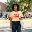 A smiling woman stands outdoors wearing a yellow tie-dye "PEACE & LOVE" t-shirt and holding a white tote bag, with food trucks and people in the background on a sunny day.