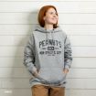A young woman with red hair wears a gray Peanuts hoodie with hands in her pockets, smiling and looking up, standing against a white wooden wall.