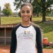 A smiling young woman stands outdoors on a baseball field, wearing a white and black Peanuts Sportswear shirt with Snoopy and a baseball graphic. Trees, benches, and sports equipment are visible in the background.