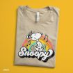 A folded beige T-shirt featuring a graphic of Snoopy dancing above a rainbow with musical notes and “Snoopy” written in bold, playful letters, displayed on a bright yellow background.
