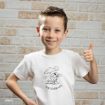 Smiling young boy with short brown hair gives a thumbs up, wearing a white t-shirt with a cartoon dog relaxing under an umbrella and the text "THE GOOD LIFE," standing in front of a light brick wall.