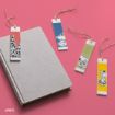Four rectangular bookmarks with Peanuts characters, each with a string, are displayed on a pink surface next to a closed gray book. The bookmarks have colorful backgrounds and cartoon illustrations.