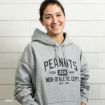 A smiling person with dark hair is wearing a gray hoodie that reads "PEANUTS 'GOOD GRIEF' NON-ATHLETIC DEPT. ESTB. 1950" and standing in front of a light wooden wall.
