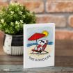 A small sign on a wooden surface shows Snoopy relaxing on a beach chair under a red umbrella, with the sun shining. The text below reads "THE GOOD LIFE." A potted plant and a brick wall are in the background.