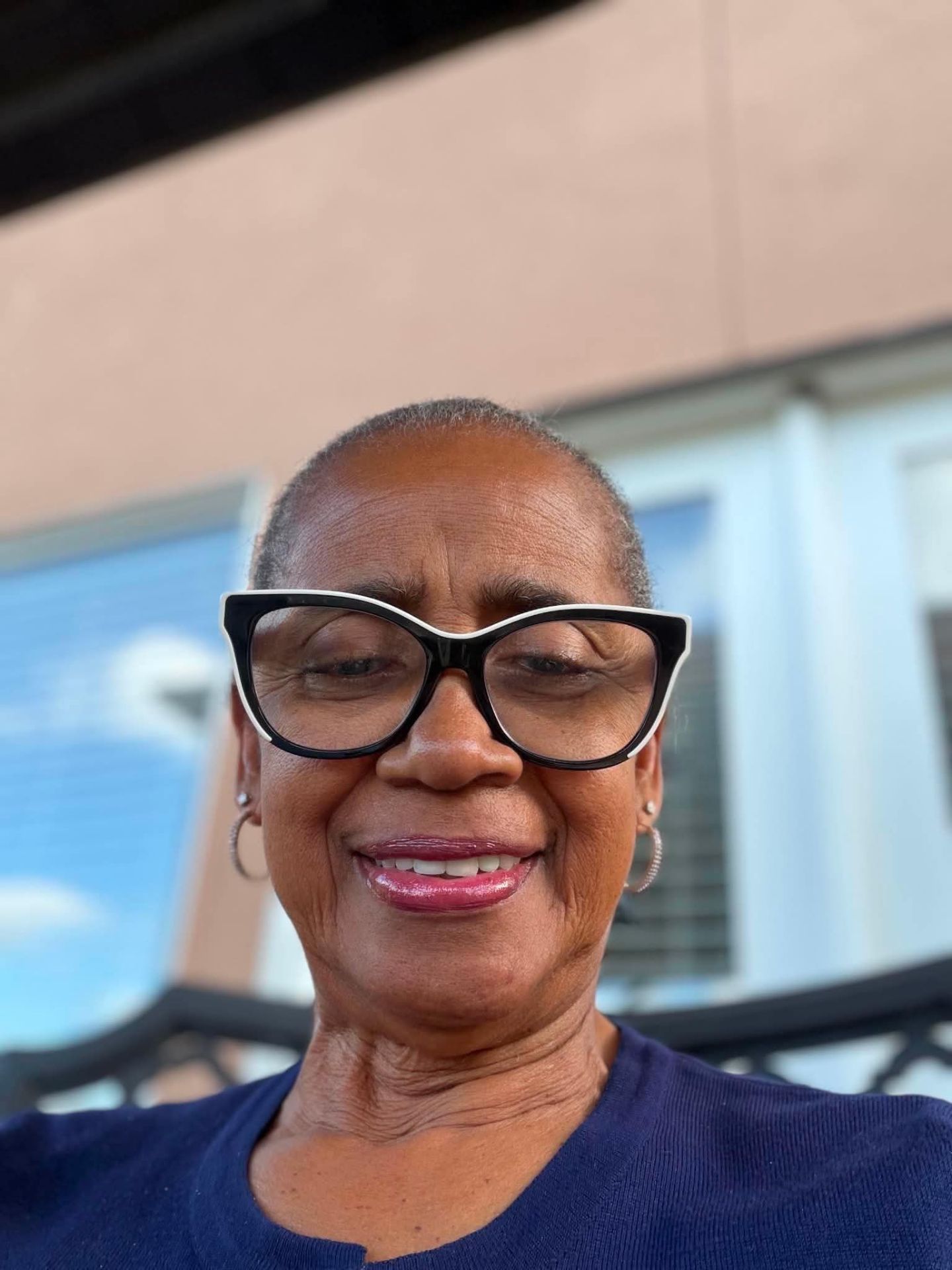 An older woman with short gray hair, large black glasses, and hoop earrings smiles at the camera. She is wearing a blue top and is seated outside in front of a building with windows.