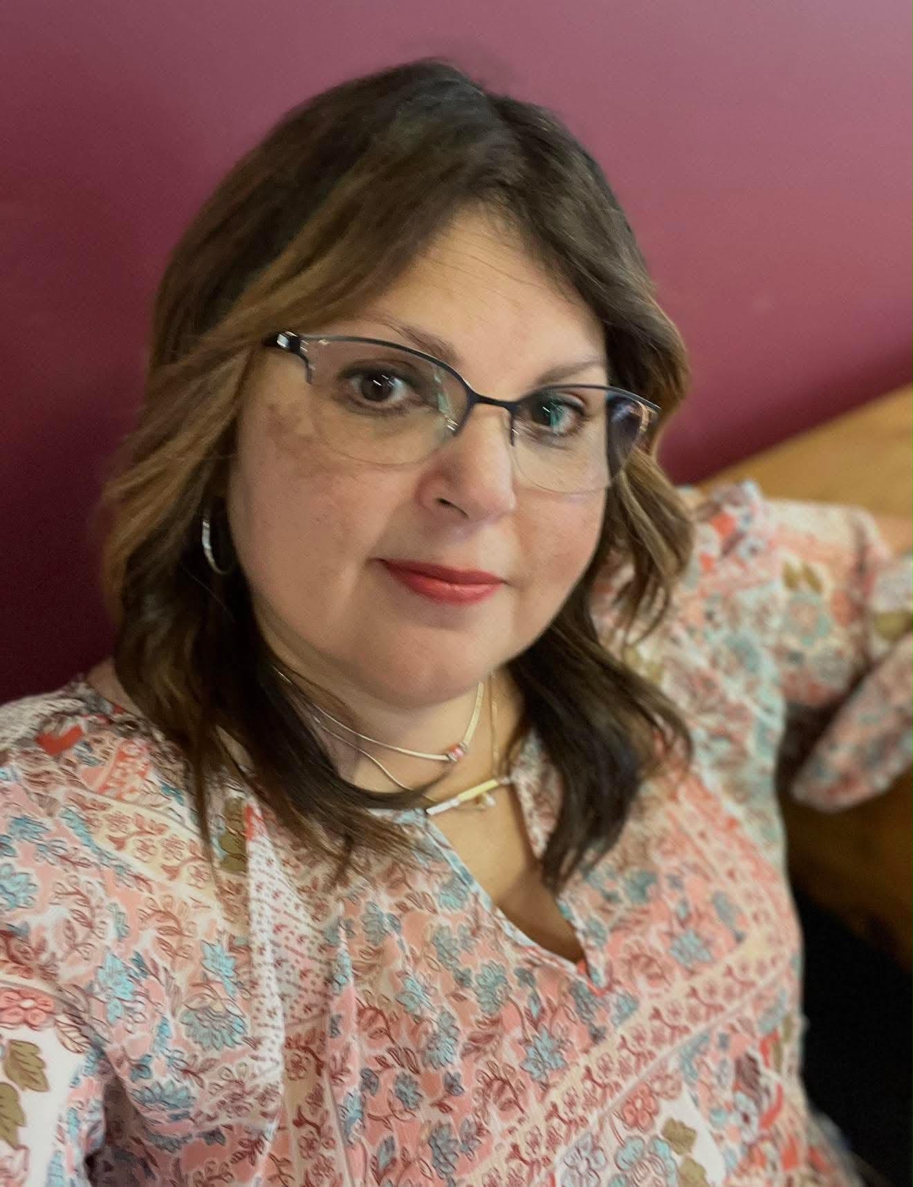 A woman with wavy brown hair and glasses is wearing a patterned blouse. She is sitting and looking at the camera with a slight smile. The background is a solid maroon color.