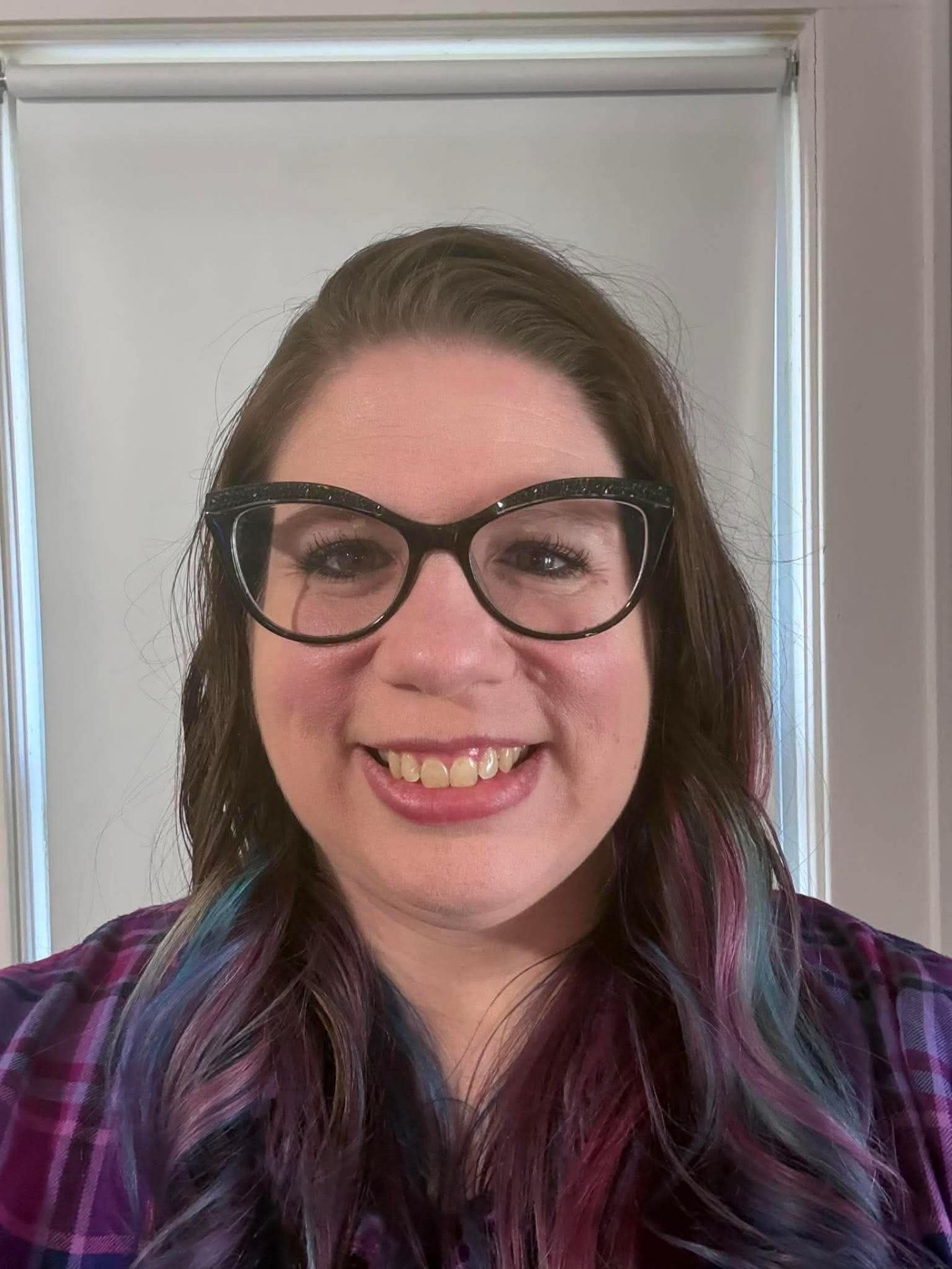 A woman with long brown hair featuring blue and purple highlights smiles at the camera. She is wearing black cat-eye glasses and a purple plaid shirt, standing indoors in front of a white window frame.