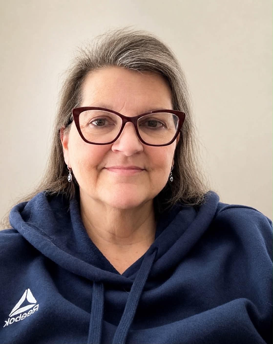 A woman with long gray hair and glasses, wearing a navy blue Reebok hoodie, smiles softly at the camera against a plain light background.