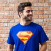 A young man with short brown hair and a beard smiles while wearing a blue T-shirt with a large red and yellow Superman logo, standing in front of a brick wall.