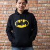A smiling young man wearing a black hoodie with a large yellow Batman logo stands in front of a brick wall, hands in the front pocket of his hoodie.
