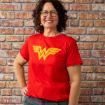 A woman with curly brown hair and glasses smiles in front of a brick wall, wearing a red T-shirt with a large yellow Wonder Woman logo on the front.
