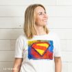 A smiling woman with blonde hair wears a white t-shirt featuring a colorful, artistic Superman logo. She stands against a white wooden wall and looks upward to her left.