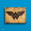 A wooden plaque with the black Wonder Woman logo is displayed against a bright blue background. The logo features stylized "W" wings, and the plaque has a slightly burnt, rustic finish.