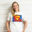 A woman with blonde hair smiles while wearing a white T-shirt featuring a colorful, artistic Superman “S” shield logo. She stands against a white wooden wall.