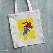 White tote bag with an illustration of Superman flying forward on a bright yellow background, his red cape flowing behind him. The Superman logo is in the bottom right corner. The bag has white handles and lies on a gray surface.