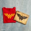 A red T-shirt with a yellow Wonder Woman logo is folded next to a wooden plaque featuring a black Wonder Woman logo, both placed on a textured light blue surface.
