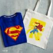 A folded blue T-shirt with a large yellow and red Superman logo next to a white tote bag featuring an illustration of Superman flying with a yellow background.