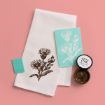 A white tea towel with a brown floral print is displayed on a pink surface, next to a turquoise floral stencil, a small turquoise eraser, and an open container of brown ink.