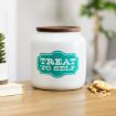 A white jar with a wooden lid sits on a table. The jar has a teal label that reads "TREAT YO SELF." Nearby are some cookies, a small plant, and a blurred green plant in the background.