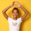 Smiling child wearing a white "Please Bring Snacks" t-shirt stands against a yellow background, holding their arms above their head and forming a heart shape with their hands.