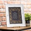 A framed print with a floral border and the quote "There are always flowers for those who want to see them" sits on a wooden table next to a small potted plant, against a brick wall background.