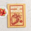 A wooden cutting board with a vintage-style design featuring a large strawberry illustration and the text "Berry Sweet, Family Fruit Fun, $1.25, Fresh Strawberries." A plate of fresh strawberries sits beside it.