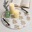 A round white tray with floral designs holds two lit pillar candles and a green glass bottle with purple flowers. An open book lies in the background on a light-colored surface.
