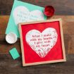 A red paper with a white heart and floral design, displaying the words "What I make with my hands, I give with my heart," is framed and placed on a wooden surface next to teal paper, paint cans, and a small teal card.