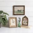 A white desk with framed art: a landscape painting on the wall, a bird illustration and a flower print on the desk, next to a green potted plant, stacked books, and a small vase with lavender.