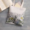A white tote bag with botanical illustrations and vintage text prints lies on a gray surface, partially filled with a closed folder. An open book is positioned nearby.